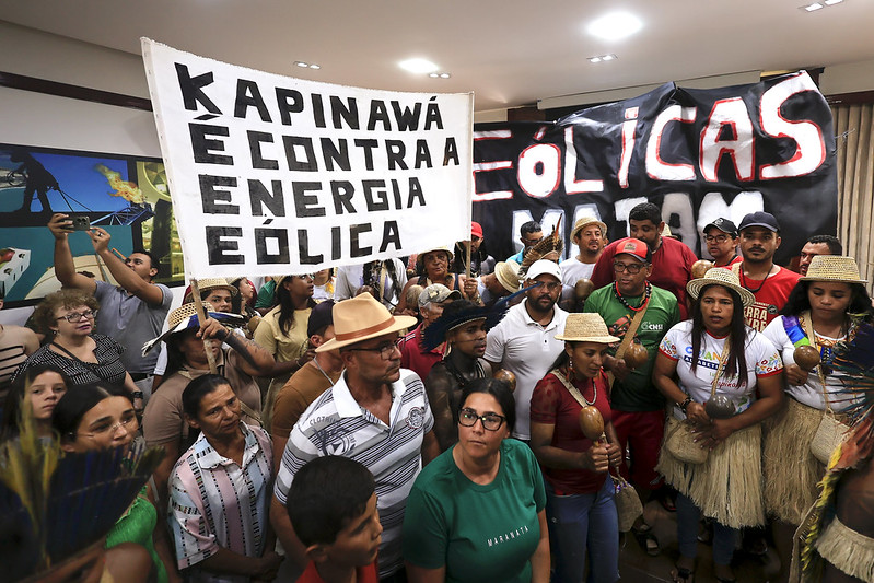 A imagem mostra um grupo de pessoas em uma sala, segurando cartazes e faixas. O cartaz principal, em letras grandes e pretas, diz KAPINAWÁ É CONTRA A ENERGIA EÓLICA. Ao fundo, há uma faixa preta com letras vermelhas e brancas que diz EÓLICAS NÃO. As pessoas estão vestidas de maneira variada, algumas com roupas tradicionais e chapéus de palha.