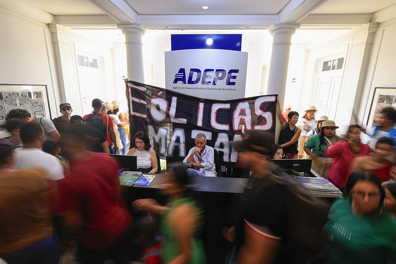 A imagem mostra um grupo de pessoas em um ambiente interno, possivelmente um escritório ou uma recepção. No centro da imagem, há uma grande faixa preta com letras vermelhas e brancas que diz EÓLICAS MATAM. Atrás da faixa, há duas pessoas sentadas em uma mesa de recepção, uma delas usando um jaleco branco. Acima da mesa, há um grande painel com o logotipo ADEPE e o texto Agência de Desenvolvimento Econômico de Pernambuco.