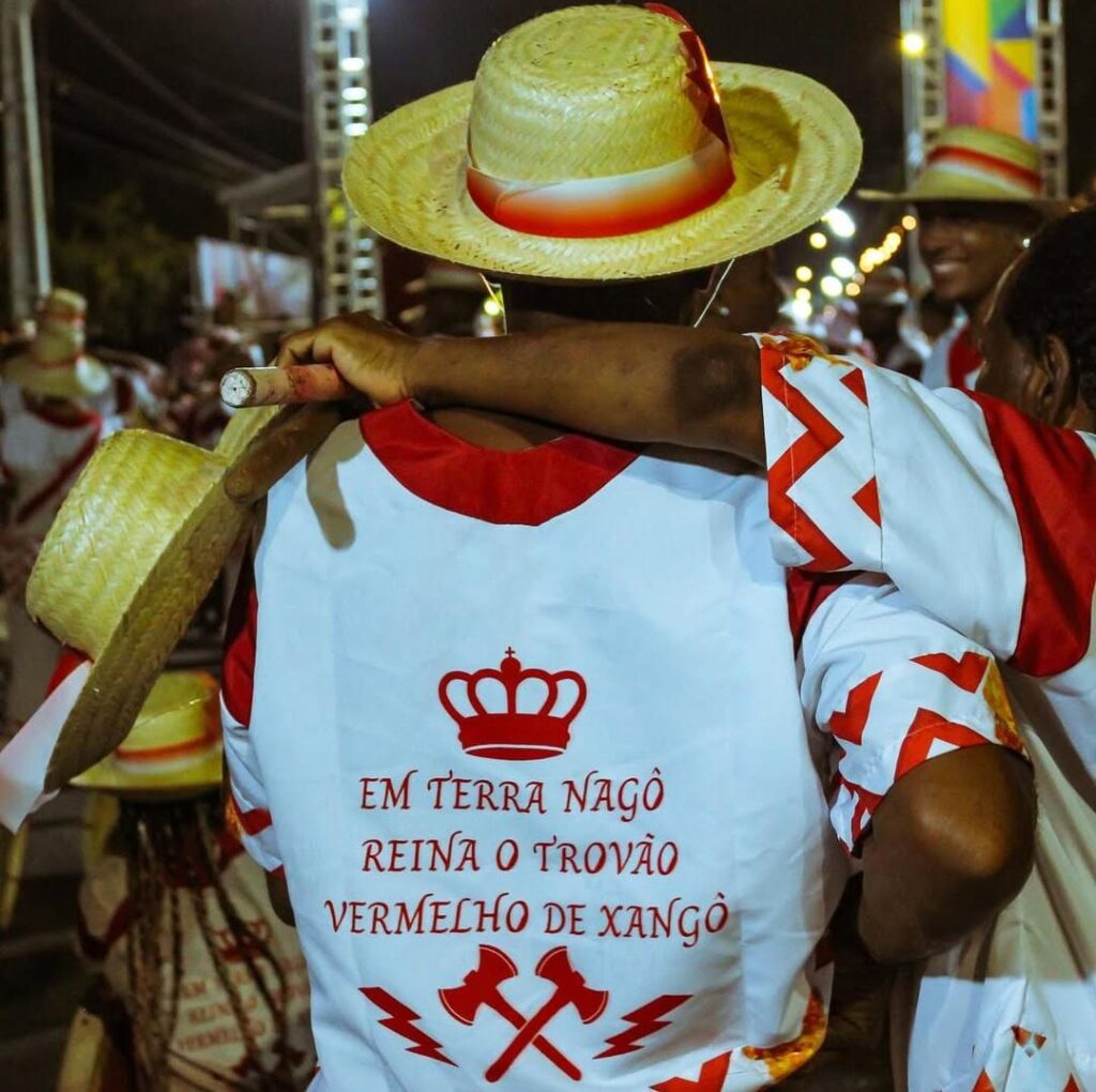 A foto mostra um homem negro vestindo uma camisa branca com detalhes vermelhos e uma frase estampada nas costas: EM TERRA NAGÔ REINA O TROVÃO VERMELHO DE XANGÔ, acompanhada de uma coroa e dois machados cruzados com raios. A pessoa usa um chapéu de palha enfeitado com uma fita vermelha e segura um instrumento de percussão no ombro. Ao redor, há outras pessoas vestindo roupas similares, algumas também com chapéus de palha, participando de um evento cultural noturno com iluminação urbana ao fundo.