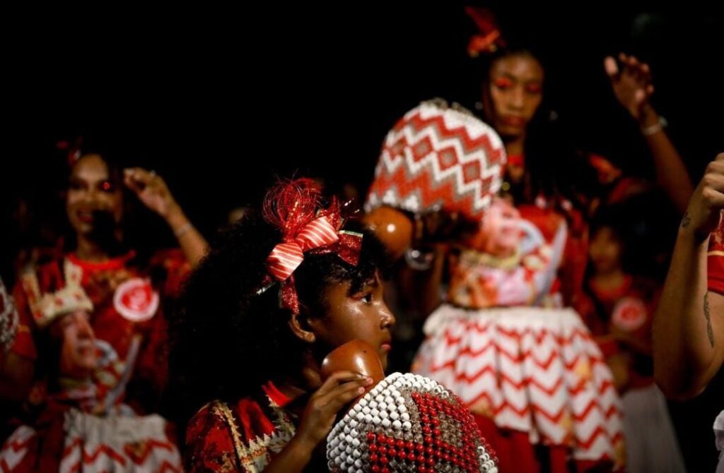 A foto mostra um grupo de pessoas vestindo roupas tradicionais coloridas, predominantemente vermelhas e brancas, com detalhes dourados. No centro da imagem, uma menina com cabelos cacheados e enfeitados com um laço vermelho segura um instrumento musical chamado xequerê, feito de cabaça com contas ao redor. Outras mulheres ao fundo também seguram o mesmo instrumento e parecem estar envolvidas em uma apresentação cultural, possivelmente ligada à música e dança afro-brasileira. A iluminação destaca os rostos e os detalhes das roupas, enquanto o fundo está mais escuro.