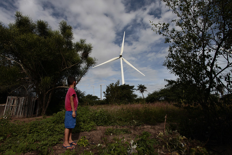 A imagem mostra um menino de pé em um ambiente rural, olhando para um grande aerogerador de energia eólica ao fundo. Ele veste uma camiseta vermelha, shorts azuis e sandálias. A paisagem ao redor é composta por vegetação, árvores e uma cerca de madeira, com um céu parcialmente nublado ao fundo.