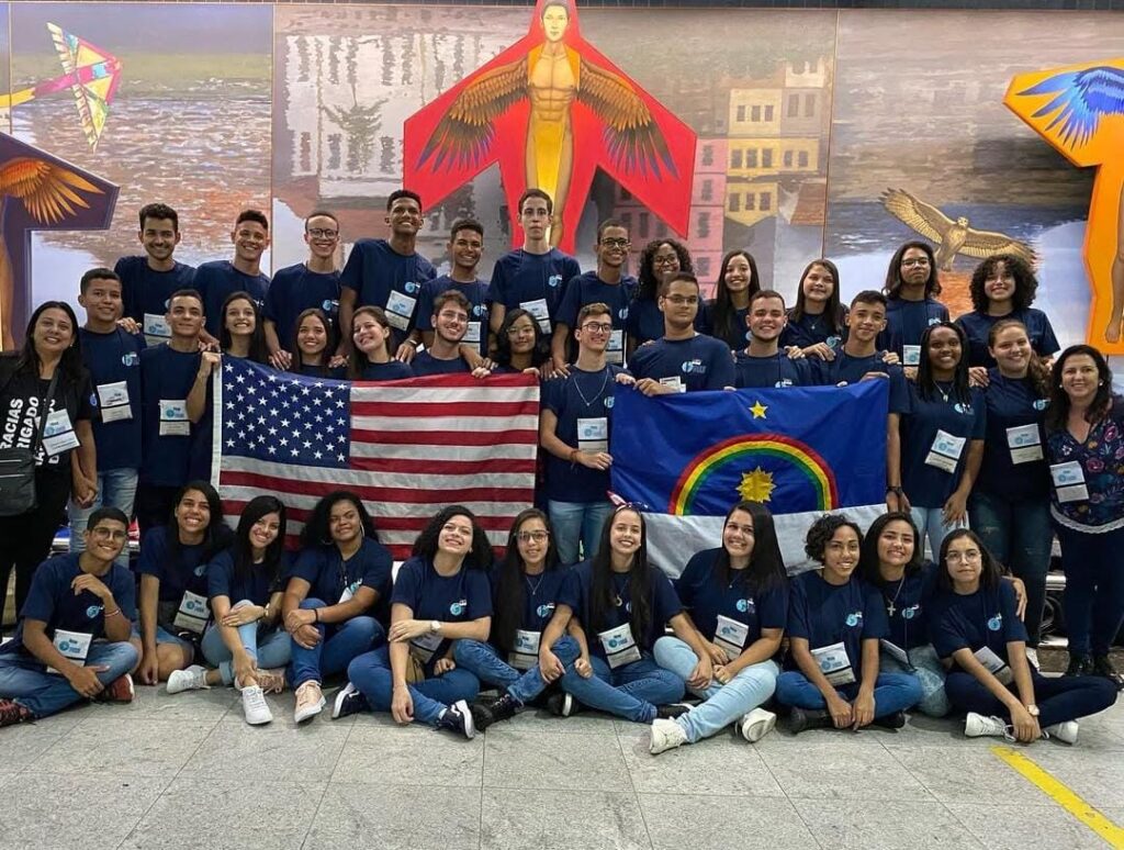 A imagem mostra um grande grupo de jovens reunidos em um ambiente fechado à frente de um mural colorido, todos vestindo camisetas azul-marinho com um logotipo no peito. Eles estão sorrindo e posando para a foto, demonstrando alegria e união. Alguns deles estão segurando duas bandeiras: a dos Estados Unidos e a do estado de Pernambuco, no Brasil.