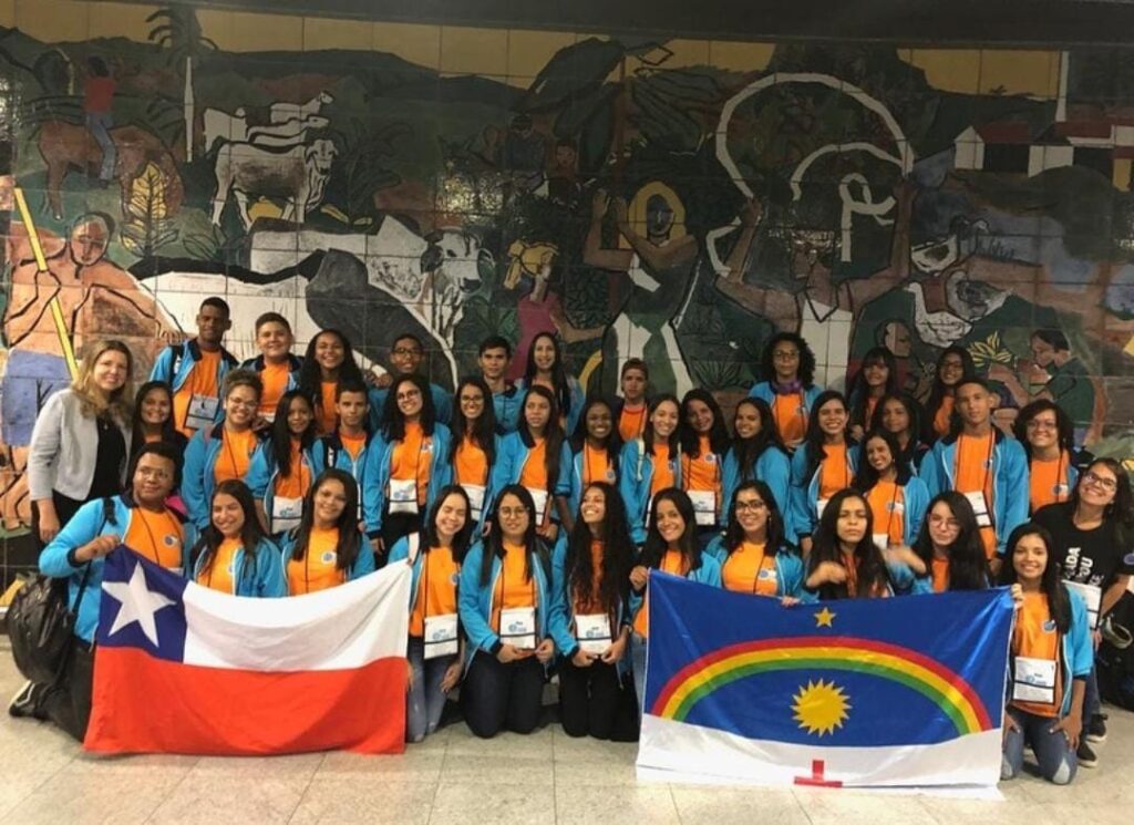 A imagem mostra um grande grupo de jovens e algumas adultas, usando camisetas laranja e casacos azul-claro, posando para uma foto. Eles parecem participar de um evento ou intercâmbio, pois usam crachás. Na frente, seguram duas bandeiras: a do Chile e a de Pernambuco. Ao fundo, um mural de azulejos retrata cenas rurais com figuras humanas, animais e elementos culturais nordestinos. O grupo exibe expressões animadas, demonstrando alegria e união.