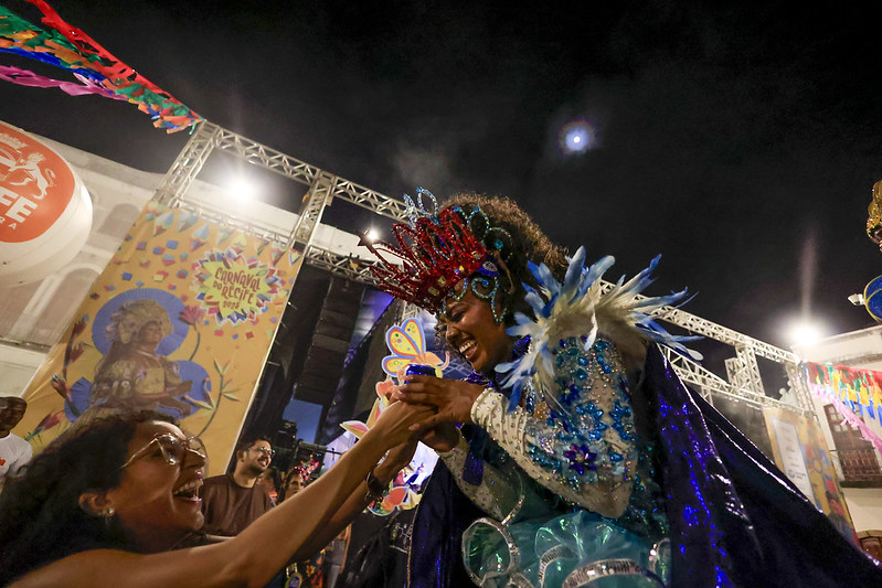 A imagem mostra uma cena vibrante do Carnaval de Recife à noite. Victoria Marques, mulher negra e jovem em uma fantasia majestosa, com penas azuis e uma coroa vermelha, sorri enquanto segura a mão de outra mulher que a admira com alegria. O fundo tem um palco iluminado, banners coloridos e a inscrição Carnaval Recife.