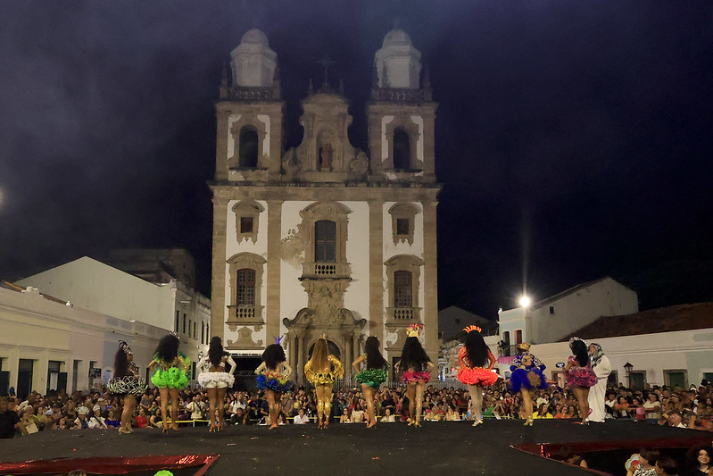 A imagem mostra um grupo de 10 mulheres em um palco, de costas para a câmera, vestidas com trajes coloridos e brilhantes. Elas estão se apresentando em frente a uma grande igreja histórica de estilo colonial, com duas torres e uma fachada ornamentada. A apresentação parece ser um evento noturno, pois o céu está escuro e há iluminação artificial. A plateia está reunida em frente ao palco, assistindo à apresentação.