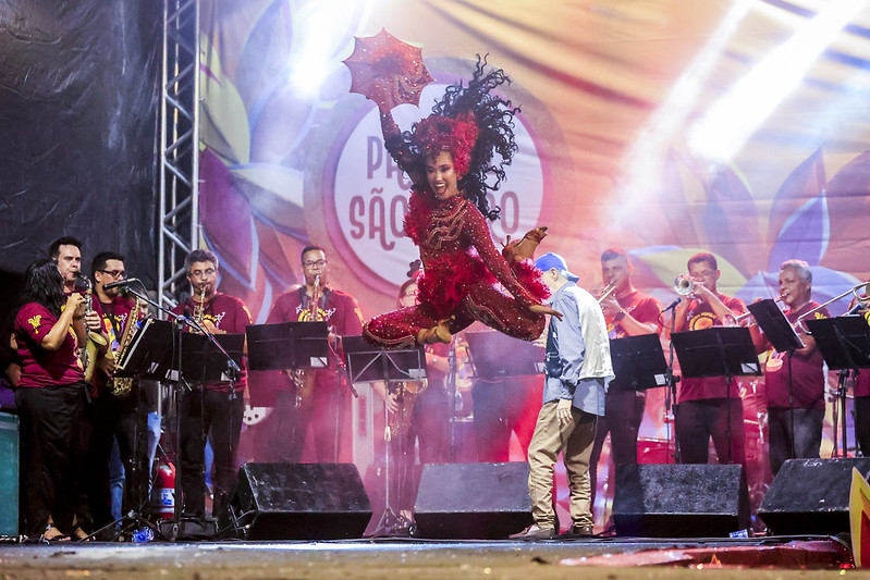 A imagem mostra uma apresentação ao vivo em um palco. No centro da imagem, Lohana Lima, mulher jovem negra, de longos cabelos pretos, vestida com uma fantasia vermelha brilhante, que parece estar no ar em um salto acrobático, segurando uma sombrinha de frevo decorada. Ao fundo há uma banda composta por várias pessoas tocando instrumentos de sopro, todas vestidas com camisetas vermelhas.