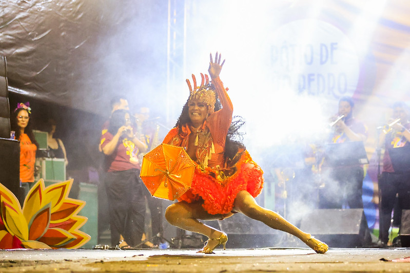 A imagem mostra Alessya Rodrigues realizando uma performance de dança em um palco. Ela está vestida com um traje vibrante e colorido, predominantemente em tons de laranja e vermelho, e segura um guarda-chuva decorativo laranja. A pessoa está em uma posição agachada, com uma perna estendida para o lado e a outra dobrada, enquanto levanta os braços para cima. Ao fundo, há uma banda tocando instrumentos de sopro e outras pessoas assistindo à apresentação. Há também uma decoração de flor grande e colorida no canto esquerdo da imagem.