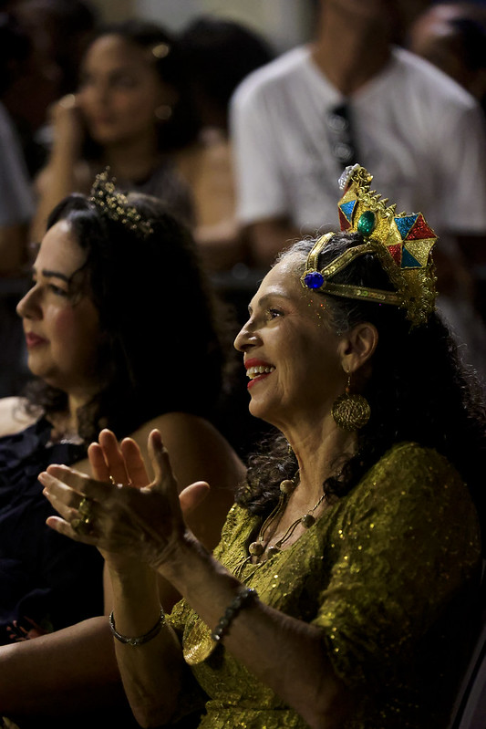 A imagem mostra duas mulheres sentadas em um ambiente festivo, que parece ser um evento ou celebração. A mulher à direita é Célia Meira, ela é idosa, branca, de longos cabelos grisalhos. Célia está vestindo uma roupa dourada brilhante e usando uma coroa colorida na cabeça similar a uma sombrinha de frevo. A pessoa à esquerda também está usando uma coroa menor, ela é mais jovem e tem cabelos negros. Ambas parecem estar aplaudindo, o que indica que estão assistindo a uma apresentação ou discurso. Ao fundo, há outras pessoas, mas estão desfocadas.