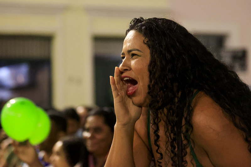 A imagem mostra uma mulher de cabelos longos, cacheados e soltos, vestindo uma roupa verde. Ela está no meio de uma multidão e parece estar torcendo ou chamando alguém, com a boca aberta e a mão próxima ao rosto, como se estivesse gritando. Seu rosto expressa emoção e entusiasmo. Ao fundo, há várias pessoas, algumas sorrindo e assistindo a algo, além de um balão verde inflado segurado por alguém na plateia.