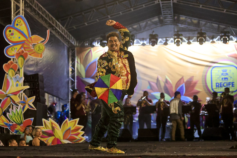 A imagem mostra uma apresentação de frevo em um palco. No centro da imagem, está Jean Carlos, um homem jovem e pardo vestido com roupas coloridas e ornamentadas, segurando uma sombrinha colorida com segmentos em vermelho, verde, amarelo e azul. A pessoa está em uma pose de dança, com um braço levantado. Ao fundo, há uma banda tocando, composta por várias pessoas, e um grande banner amarelo com flores coloridas e o nome RECIFE escrito em azul. O palco é decorado com grandes figuras de borboletas e flores coloridas. A iluminação do palco é intensa, destacando a apresentação.