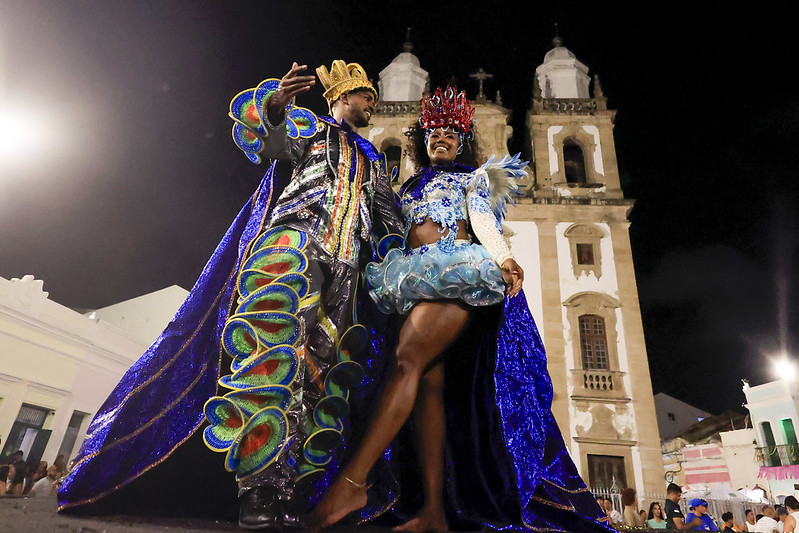 A imagem mostra um casal majestoso vestido com trajes luxuosos e coloridos em uma celebração noturna. O homem, representando um rei, usa uma coroa dourada e uma capa azul brilhante com detalhes coloridos e ondulados. Sua roupa é cheia de brilhos e bordados sofisticados. A mulher, representando uma rainha, veste uma fantasia azul clara e brilhante, com penas e detalhes exuberantes. Ela usa uma coroa vermelha adornada com pedras e brilhos. Eles posam com confiança e alegria, olhando para o público ou para algo ao redor. Ao fundo, há uma imponente igreja colonial de fachada branca com duas torres, iluminada pela luz artificial da cidade.