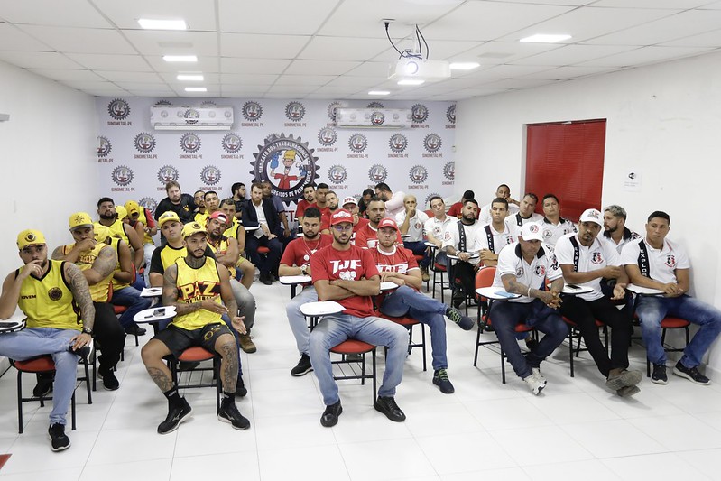 A imagem mostra uma sala de aula ou pequeno auditório de paredes claras com várias homens jovens sentados em cadeiras com pranchetas. Os homens são integrantes de torcidas organizadas do Sport Recife, Náutico e Santa Cruz e estão vestidos com camisetas de cores variadas, principalmente amarelas, vermelhas e brancas, e algumas delas têm bonés. No fundo da sala, há um banner com o logotipo de uma organização sindical. A sala tem um teto com iluminação embutida e um projetor pendurado. As pessoas estão olhando para a frente, como se estivessem assistindo a uma apresentação ou aula.