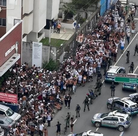 A imagem é uma vista aérea de um grande número de pessoas aglomeradas em uma calçada, cercadas por vários policiais e viaturas. As pessoas parecem estar sem camisa e algumas estão com as mãos para cima. Há uma cerca metálica ao lado da calçada e, do outro lado da cerca, há um prédio com uma área verde. Na rua, há várias viaturas policiais estacionadas, e alguns policiais estão em pé, observando a multidão. A cena parece ser de uma operação policial ou uma manifestação.