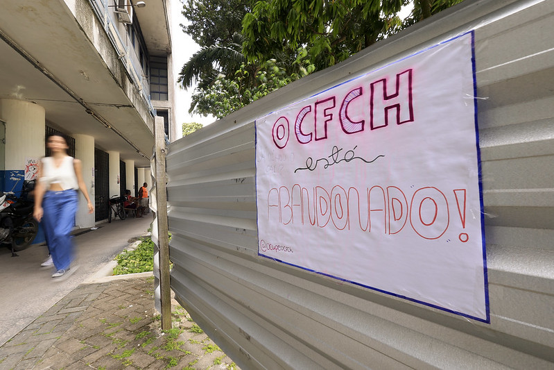 A imagem mostra uma área externa de um prédio. À direita, há uma cerca de metal cinza e à esquerda, uma calçada. Na cerca, há um cartaz branco com bordas azuis e letras grandes em vermelho e rosa que diz: O CFCH está do lado!. Ao fundo, há uma pessoa caminhando, parcialmente desfocada, e outras pessoas mais distantes. A imagem parece ser de um ambiente universitário ou escolar, com árvores e vegetação ao fundo.