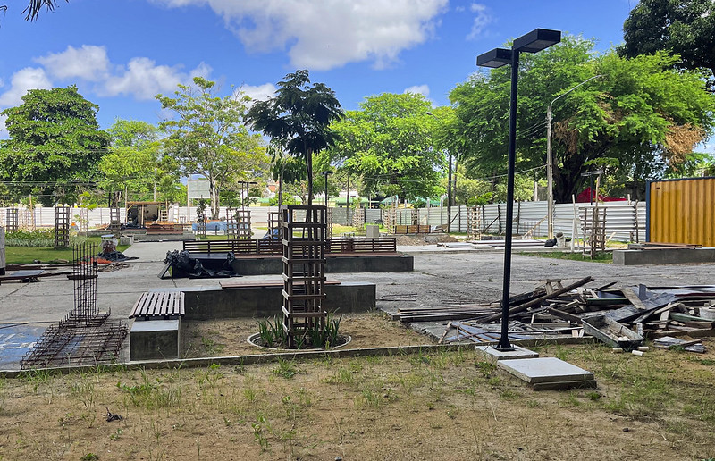 A imagem mostra um canteiro de obras ao ar livre, possivelmente em um parque ou praça. Há várias estruturas de metal em construção, algumas ao redor de árvores jovens. O chão é pavimentado com blocos de concreto, e há bancos de madeira espalhados pelo local. À direita, há um poste de iluminação e uma área com materiais de construção, incluindo pedaços de madeira e metal. Ao fundo, há uma cerca branca e árvores verdes, sugerindo que o local está em uma área arborizada. O céu está azul com algumas nuvens brancas.