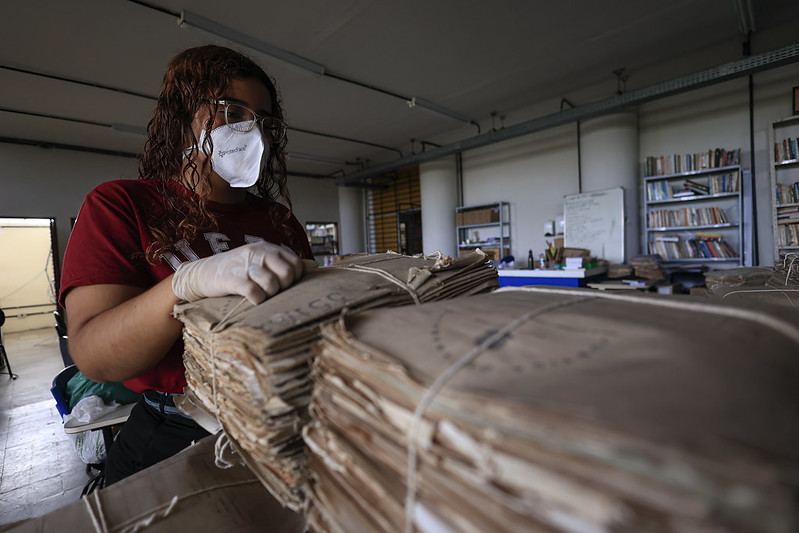 A imagem mostra uma pessoa em um ambiente interno, possivelmente um arquivo ou biblioteca, manuseando um grande volume de documentos ou jornais empilhados e amarrados com barbante. A pessoa está usando luvas brancas e uma máscara facial, sugerindo que está lidando com materiais que precisam ser manuseados com cuidado, talvez por serem antigos ou frágeis. Ao fundo, há estantes com livros e outros materiais organizados.