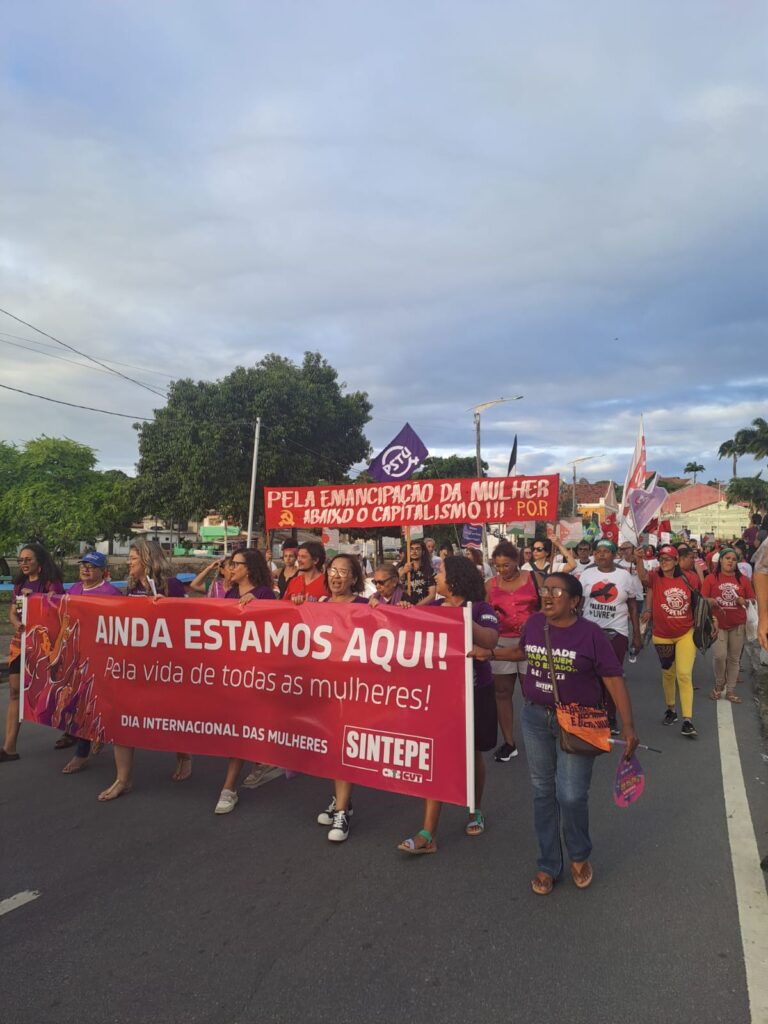 A foto mostra uma marcha do Dia Internacional das Mulheres, com um grupo segurando uma faixa vermelha que diz AINDA ESTAMOS AQUI! Pela vida de todas as mulheres!. Atrás, outra faixa vermelha defende a emancipação feminina. Muitas pessoas participam, vestindo roxo e vermelho, segurando bandeiras e cartazes. O protesto ocorre em uma rua, com céu parcialmente nublado e árvores ao fundo.