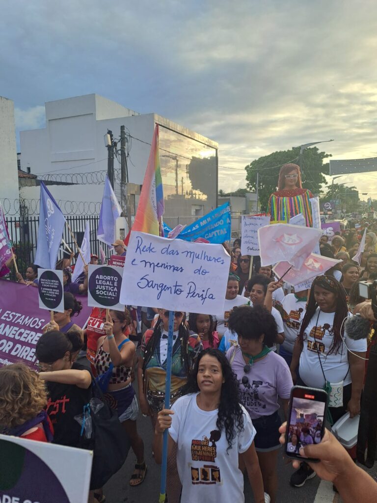 A foto mostra uma marcha do Dia Internacional das Mulheres com diversas participantes segurando cartazes e bandeiras. No centro, uma mulher segura um cartaz escrito Rede das mulheres e meninas do Sargento Perifa. Outras pessoas carregam placas com mensagens como Aborto legal e justiça social. Há bandeiras coloridas e uma boneca gigante representando uma mulher com roupas coloridas. A manifestação ocorre ao ar livre, com céu parcialmente nublado e um grande grupo reunido.