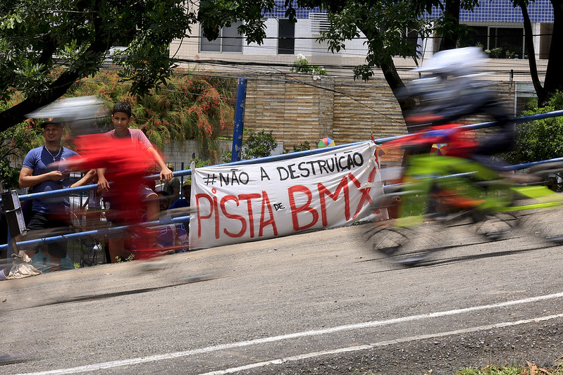 A imagem mostra uma pista de BMX com ciclistas passando rapidamente, criando um efeito de desfoque devido à velocidade. Ao fundo, há um cartaz branco com letras pretas e vermelhas que diz: #NÃO À DESTRUIÇÃO DA PISTA DE BMX. Duas pessoas estão em pé ao lado do cartaz, observando a pista. A cena parece ser de um evento ou manifestação em defesa da pista de BMX, destacando a importância de preservá-la.