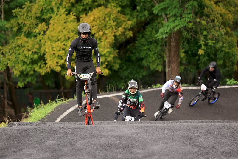 A imagem mostra uma competição de ciclismo BMX em uma pista ao ar livre. Quatro ciclistas estão na pista, todos usando capacetes e roupas de proteção. O ciclista na frente, com o número 102, está fazendo uma manobra chamada empinada, onde a bicicleta fica apenas na roda traseira. Atrás dele, três ciclistas com os números 32, 51 e 6 seguem em diferentes posições na pista. A pista é asfaltada, com curvas e elevações. Ao fundo, árvores com folhas verdes e amarelas