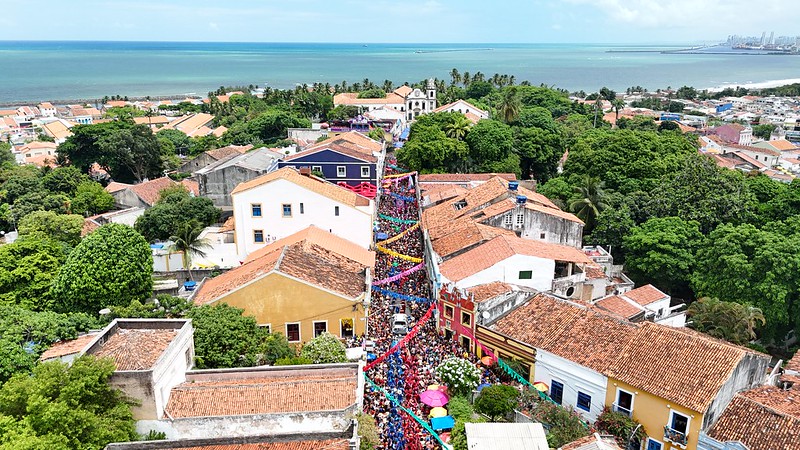 A imagem mostra uma vista aérea do sítio histórico de Olinda durante o carnaval. No centro da imagem, há uma rua estreita cheia de pessoas, decorada com bandeirinhas coloridas que atravessam de um lado ao outro. As casas ao longo da rua têm telhados de telha vermelha e fachadas coloridas, principalmente em tons de amarelo, branco e vermelho. Ao fundo, é possível ver o mar e uma faixa de vegetação densa.