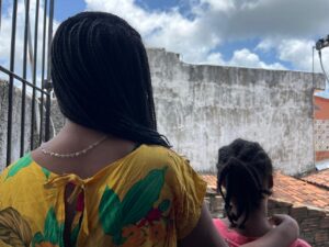 A imagem mostra uma mullher e uma menina negras vistas de costas. A pessoa à esquerda tem cabelos trançados e usa uma blusa amarela com detalhes verdes e vermelhos. A pessoa à direita é uma criança com cabelos presos em pequenos coques. Ambas estão olhando para uma parede de concreto desgastada, com um céu azul e algumas nuvens ao fundo. A pessoa à esquerda está usando um colar prateado. A imagem parece ser tirada em um ambiente externo, possivelmente em um quintal ou varanda.