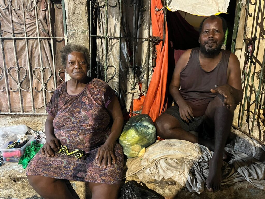 A imagem mostra um homem e uma mulher sentados em frente a um abrigo improvisado, com grades e tecidos pendurados. A mulher, de cabelos curtos e grisalhos, veste um vestido estampado e tem expressão séria. O homem, com barba e regata escura, parece gesticular enquanto fala. O ambiente sugere vulnerabilidade social, com sacolas, roupas e objetos espalhados. A iluminação artificial destaca os contrastes de luz e sombra no local.