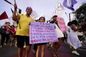 A foto mostra uma manifestação feminista em uma rua, possivelmente no Dia Internacional da Mulher. No centro, uma menina segura um cartaz roxo denunciando o feminicídio de 10.000 mulheres em 9 anos. Ao lado dela, uma mulher de blusa amarela e lenço lilás segura um pedaço de madeira. Ao fundo, várias manifestantes carregam cartazes e bandeiras com mensagens contra a violência de gênero. Uma delas segura um cartaz rosa com as hashtags e #PAREMDEMATAR.