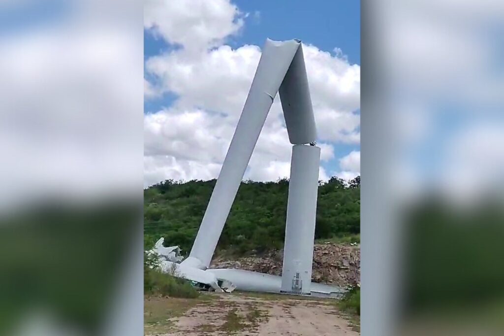 Imagem de aerogerador de parque eólico dobrado ao meio e caído em uma área rural, ao lado de uma estrada de terra.