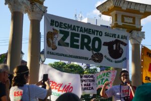 A foto mostra uma manifestação ao ar livre durante o dia, com céu azul e sol. Pessoas estão reunidas em frente a uma estrutura de colunas antigas. Uma faixa grande pede Desmatamento Zero e Salve a APA Aldeia-Beberibe, com desenhos de animais como onça, cobra e preguiça. Outras faixas menores defendem a natureza e a justiça social. Um homem com camiseta branca e microfone lidera o protesto. Algumas pessoas tiram fotos e acompanham atentas. O clima é pacífico e de engajamento ambiental. A manifestação pede a proteção da floresta da APA Aldeia-Beberibe.