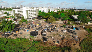 A imagem mostra uma área aberta com diversas construções improvisadas, que parecem ser barracos ou abrigos temporários feitos de materiais como madeira, plástico e lonas. Elas estão espalhadas de forma desorganizada sobre um terreno de terra, cercado por vegetação rasteira. Ao fundo, há um contraste marcante: edifícios residenciais brancos de vários andares e, mais ao longe, uma cidade com muitos prédios altos. A perspectiva aérea da foto permite visualizar tanto essa área de habitação precária quanto a cidade ao fundo.