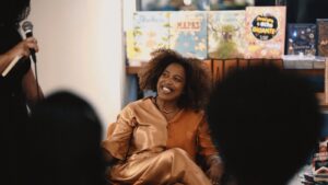 Essa imagem mostra uma cena de um evento, possivelmente uma roda de conversa ou lançamento de livro. Em destaque, Midia Noelle, mulher negra e jovem está sentada confortavelmente em uma poltrona. Ela está sorrindo com alegria e atenção, olhando para alguém fora do quadro que segura um microfone — provavelmente uma entrevistadora ou mediadora da conversa. Ela usa uma roupa de cetim brilhante em tom cobre, com blusa e calça combinando. Ela tem cabelo cacheado, volumoso e solto, e usa colares finos e discretos. Atrás dela, há uma estante com vários livros coloridos expostos, alguns com capas ilustradas voltadas para o público.