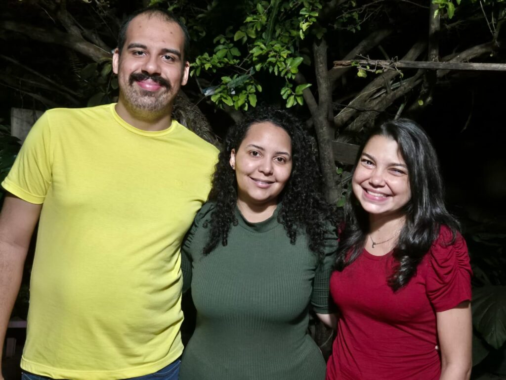 A imagem mostra três pessoas em pé, lado a lado, em uma foto noturna. Da esquerda para a direita, está Wander Vieira; ele é um homem alto com camisa amarela; no meio está Josyclaudia Gomes. uma mulher com uma blusa verde e ao seu lado outra mulher, Elismarcia Tosta, vestida com blusa vermelha, ambas de estatura média e ao fundo mostra uma pequena árvore.