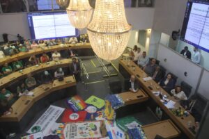 A imagem captura uma reunião em uma sala ampla e bem iluminada, que parece estar ocorrendo em um contexto oficial ou público. As pessoas estão dispostas em torno de uma mesa em formato de U, com alguns participantes usando bonés verdes e camisetas com logotipos, aparentemente representando organizações ou movimentos. Há faixas e bandeiras coloridas espalhadas no centro da sala, com mensagens e logotipos diversificados. Nas paredes ao fundo, duas grandes telas de projeção exibem slides cujo tema central é a Síndrome da Turbina Eólica (STE). O texto na tela menciona estudos sobre os impactos de turbinas eólicas na saúde, destacando sintomas como desequilíbrio, dores de cabeça, problemas auditivos e de sono, associados ao ruído de baixa frequência e infrassons dessas turbinas. O teto exibe dois lustres de cristal, acrescentando um toque sofisticado ao espaço. A cena transmite a ideia de uma audiência pública ou uma reunião de trabalho para discutir questões relacionadas ao tema apresentado.