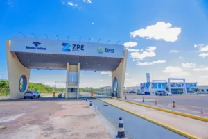 Esta é uma imagem da entrada de uma Zona de Processamento de Exportação (ZPE) localizada em Parnaíba, no Piauí, Brasil. No centro, há um grande pórtico com um design moderno, onde está escrito ZPE Parnaíba - PI. Também estão visíveis os logotipos da Receita Federal e do Governo do Estado do Piauí. À direita, vemos um edifício de fachada colorida e moderna, com alguns carros estacionados na frente. O céu é claro e com poucas nuvens, sugerindo um dia ensolarado. A estrutura reflete uma área voltada para atividades industriais e de exportação.