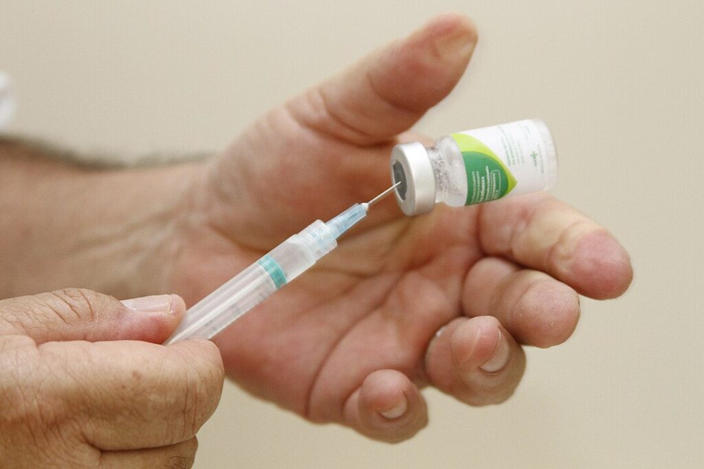 Foto de uma mão humana segurando um pequeno vidro de vacina com rótulo branco e verde, enquatro outra mão segura a seringa usada para extrair o liquido de dentro do recipiente.