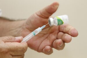 Foto de uma mão humana segurando um pequeno vidro de vacina com rótulo branco e verde, enquatro outra mão segura a seringa usada para extrair o liquido de dentro do recipiente.