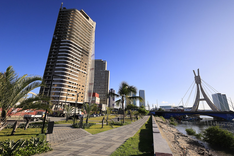 A imagem mostra uma cidade costeira com um grande corpo de água à direita e uma ponte conectando duas partes da cidade. Veículos circulam sobre a ponte, destacando sua importância para a mobilidade. À direita, há vários edifícios modernos, incluindo um arranha-céu em construção. No fundo, a cidade se estende com casas e prédios menores, misturando áreas residenciais e comerciais. Há espaços verdes com pequenas palmeiras e uma praça próximo à água.