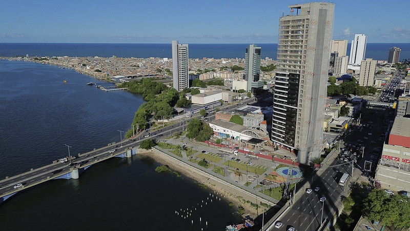 A imagem mostra o bairro do Pina, no Recife, uma área urbana costeira com uma grande ponte conectando suas duas partes. À esquerda, há um corpo de água, um rio, com o mar ao fundo. À direita, a área urbana tem vários prédios altos, incluindo um em construção. A cidade mistura arquitetura moderna com espaços verdes. Há árvores e um parque próximo à água, criando um contraste harmonioso. Ao fundo, vê-se casas e prédios menores. A foto foi feita durante dia claro, com céu azul e poucas nuvens.