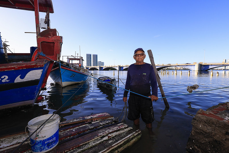 Nesta foto, vemos um homem idoso, com pele morena e barba branca rala, em pé dentro d'água até os joelhos, ao lado de barcos de pesca atracados. Ele veste uma camisa de manga longa azul escura, bermuda preta e usa boné e óculos escuros. Ele segura uma corda presa a um dos barcos. O cenário é um porto ou cais simples, com um céu claro e azul ao fundo. Atrás do homem, há uma ponte grande que cruza um rio. Ao fundo, também é possível ver alguns prédios altos de uma cidade grande, o Recife. À frente dele, há um pequeno trapiche de madeira desgastada e um balde branco, aparentemente cheio d’água.