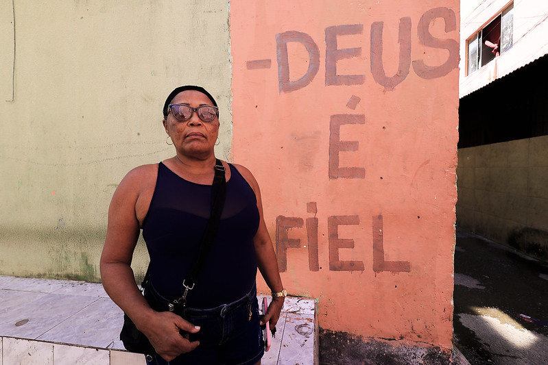 Foto de Walquíria Dias. Ela é uma mulher negra, de cabelos presos na nuca, usando óculos escuros e uma bolsa de alça preta cruzada sobre a blusa da mesma cor. Na mão esquerda, ela segura um telefone celular. Walquíria foi fotograda em ambiente externo, à frente de uma parede pintada de rosa com a frase “Deus é fiel” pintada em letras vermelhas.