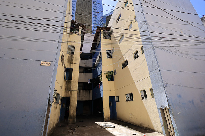 A imagem mostra um conjunto de prédios residenciais pintados de amarelo e azul, com vários fios elétricos cruzando a cena na frente dos edifícios. As construções possuem janelas pequenas protegidas por grades e algumas plantas penduradas, trazendo um pouco de verde ao ambiente. Na parede à esquerda, há uma placa escrita BLOCO J. O chão entre os prédios está sujo e há uma escada que leva ao interior dos edifícios. Ao fundo, destacando-se no horizonte, há um prédio alto e moderno, contrastando com as construções em primeiro plano.
