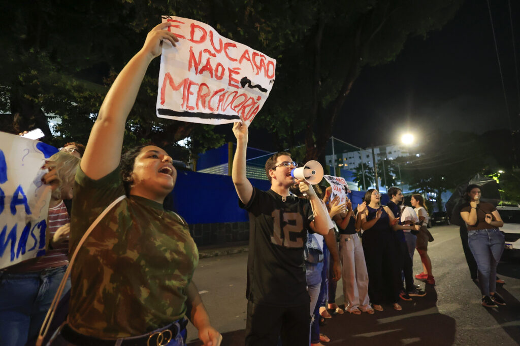 A foto mostra um grupo de estudantes protestando à noite, em uma rua iluminada. Uma jovem em destaque segura um cartaz escrito “Educação não é mercadoria” e grita palavras de ordem. Ao lado dela, um rapaz fala em um megafone. Outras pessoas atrás também seguram cartazes e demonstram indignação. O clima é de luta e resistência contra problemas no ensino superior privado.