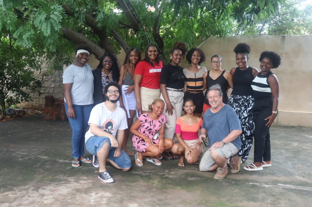 Foto de um grupo de 13 pessoas reunidas em um espaço externo, sob uma árvore, com um muro ao fundo. Nove estão em pé e quatro agachadas à frente. A maioria são mulheres negras jovens, com diferentes penteados afro e roupas casuais. Dois homens brancos estão na frente, agachados, sorrindo. O ambiente é simples, com chão de cimento e vegetação ao redor. A expressão de todos transmite alegria, acolhimento e união. A imagem sugere um encontro cultural, educativo ou comunitário.