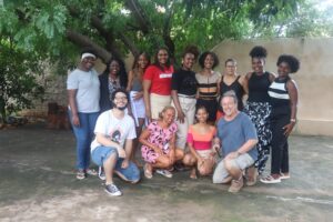 Foto de um grupo de 13 pessoas reunidas em um espaço externo, sob uma árvore, com um muro ao fundo. Nove estão em pé e quatro agachadas à frente. A maioria são mulheres negras jovens, com diferentes penteados afro e roupas casuais. Dois homens brancos estão na frente, agachados, sorrindo. O ambiente é simples, com chão de cimento e vegetação ao redor. A expressão de todos transmite alegria, acolhimento e união. A imagem sugere um encontro cultural, educativo ou comunitário.