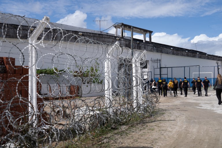 A imagem mostra a fachada do presídio do Curado, em Pernambuco, uma área cercada por arame farpado enrolado em espirais ao redor de uma cerca branca de metal. Atrás da cerca, o prédio é branco com um telhado escuro e algumas antenas no topo. No lado direito, um grupo de pessoas vestidas com camisetas pretas e amarelas caminha em direção ao presídio. O chão é de terra e há algumas plantas crescendo perto da cerca. O céu está parcialmente nublado, com algumas nuvens brancas.