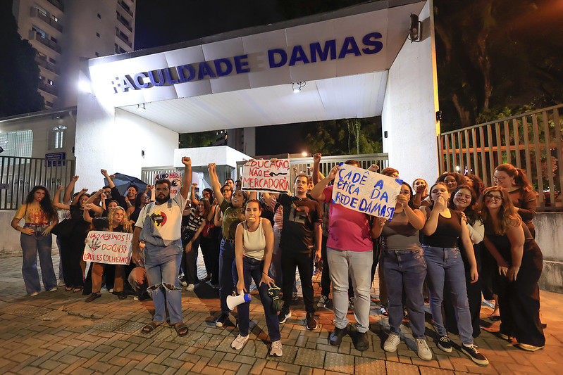 A foto mostra um grupo de pessoas reunidas em frente ao portão da Faculdade Damas, durante a noite. Elas estão participando de um protesto e seguram cartazes e faixas com mensagens de reivindicação. Alguns dos cartazes contêm frases como Educação não é mercadoria, Cadê a ética da rede Damas? e Acadêmicos de Damas unidos e lutando. Muitas das pessoas estão com os braços erguidos e algumas seguram megafones, demonstrando energia, determinação e engajamento na causa.