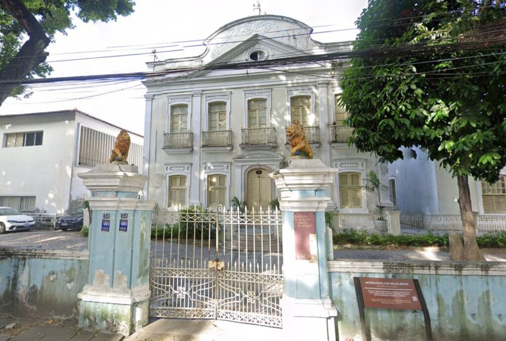 Foto da fachada da Facepe, um edifício histórico com uma arquitetura clássica e sofisticada. Ele tem dois andares e é pintado de branco, com detalhes ornamentais em alto-relevo que decoram sua fachada. As janelas possuem molduras elegantes e sacadas com grades de ferro, contribuindo para um ar refinado. Na entrada há um portão de ferro, sustentado por dois pilares altos. No topo de cada um deles repousa uma imponente estátua dourada de leão, conferindo um aspecto de grandiosidade e prestígio ao local. À esquerda do edifício principal, é possível ver um prédio moderno, também branco, contrastando com a estética clássica da construção histórica. Do lado direito, há uma grande árvore de folhagem verde, trazendo um toque natural ao ambiente.
