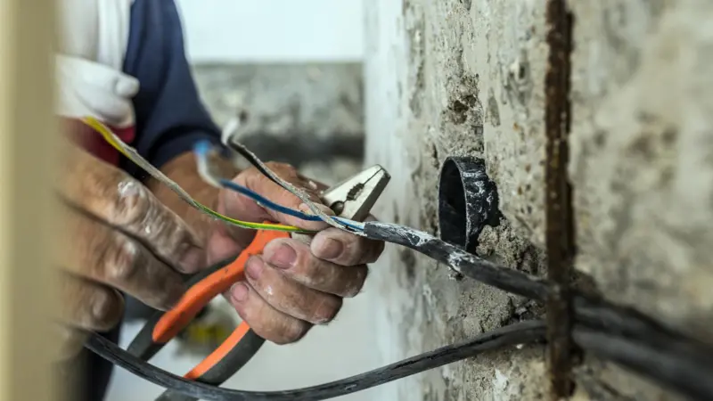 A imagem mostra as mãos de um eletricista trabalhando em uma instalação elétrica em uma parede de concreto. Ele segura fios coloridos com um alicate, enquanto realiza algum tipo de reparo ou conexão. A parede está parcialmente descascada, revelando os conduítes por onde passam os cabos. O ambiente parece estar em reforma ou construção.
