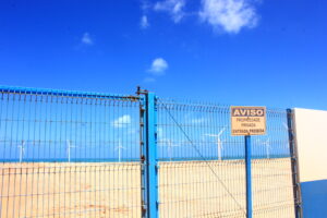 A foto mostra uma cerca de metal pintada de azul, que bloqueia o acesso a uma área de praia. No centro da cerca há um portão trancado com uma corrente. À direita do portão, há uma placa de aviso afixada na grade. A placa está escrita em português e diz: AVISO PROPRIEDADE PRIVADA ENTRADA PROIBIDA. Atrás da cerca, no fundo da imagem, pode-se ver o mar azul e várias turbinas eólicas brancas (aquelas grandes hélices usadas para gerar energia com o vento). O céu está limpo e muito azul, com algumas poucas nuvens brancas.